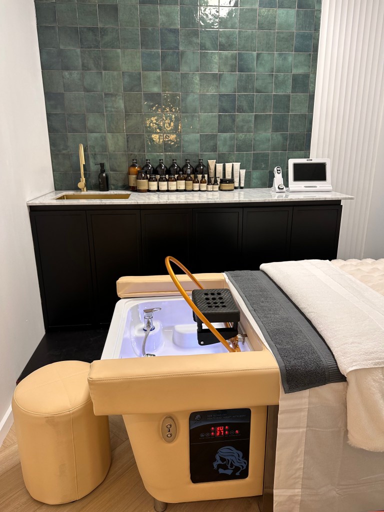 Interior of a spa treatment room featuring a modern treatment table with a built-in basin and digital display, surrounded by green tiled walls and a collection of skincare products on a countertop.